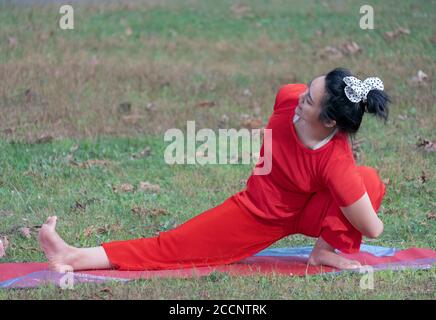 Eine Frau, die einen roten Hosenanzug und einen Haarbogen mit Polka-Punkt trägt, macht einen schwierigen Stretcn bei einem Yoga-Kurs in einem Park in Queens, New York Stockfoto