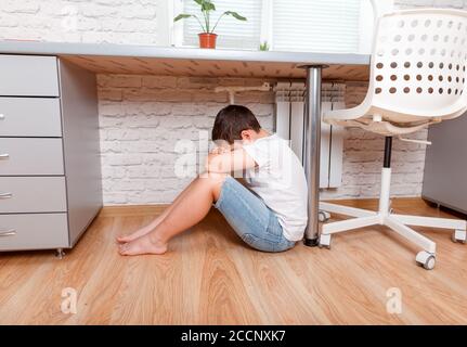 Vorteen Junge sitzt unter Schreibtisch, bedeckt sein Gesicht mit Händen zu Hause Stockfoto