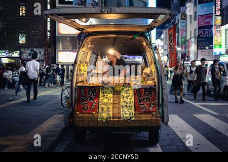 Yakitori-Stand: Frau, die nachts in einem Van Spieße kocht und verkauft. Leute, die auf der Straße gehen, essen Street Food. Shinjuku, Tokio, Japan Stockfoto