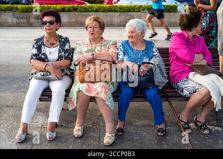 Frontansicht von vier spanischen Rentnerinnen der Mittelklasse, die auf einer Holzbank sitzen und sich unterhalten und das Leben in Paseo Pereda, Santander, Spanien, vorbeiziehen sehen. Stockfoto