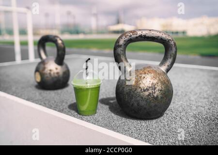 Gesundheit und Fitness Green Smoothie Detox Drink im Fitnessstudio mit Kettlebells Gewichten im Outdoor Training Fitness Center. Plastikbecher Gemüsesaft Stockfoto