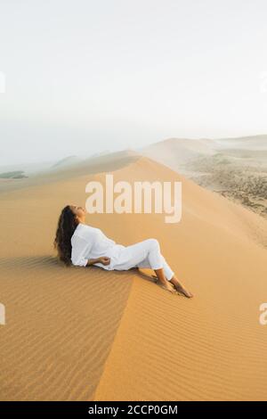 Schöne Brünette Frau in weißen Kleidern auf Sanddüne in Marokko Sahara Wüste entspannen. Nebel am Morgen vor Sonnenaufgang. Harmonie mit der Natur. Stockfoto