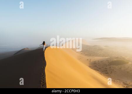 Frau, die auf einer riesigen Sanddüne in der Sahara von Marokko läuft. Schönes warmes Sonnenlicht und Nebel am Morgen. Blick von hinten. Freiheitskonzept. Stockfoto