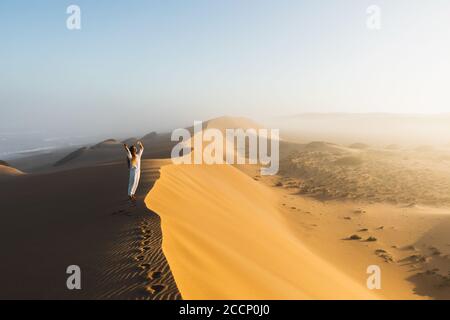 Frau, die den Sonnenaufgang auf einer riesigen Sanddüne genießt. Schönes warmes Sonnenlicht und Nebel am Morgen. Sahara-Wüste, Marokko. Stockfoto