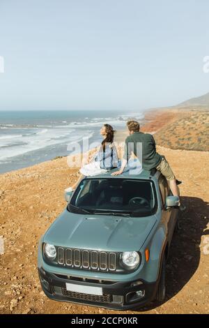 Roadtrip-Konzept. Junges und glückliches Hippie-Paar, das auf dem Dachauto sitzt und einen fantastischen Blick auf das Meer und die Küste Marokkos genießt. Fernweh und Lifestyle. Stockfoto