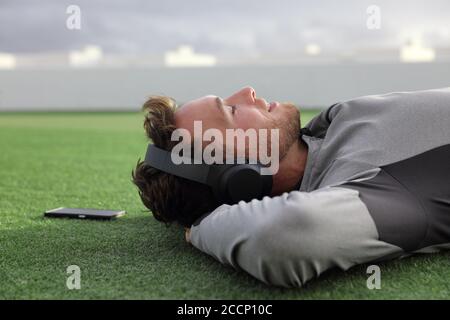 Glücklicher Mann, der mit Kopfhörern Telefonmusik hört, entspannt schläft oder meditiert auf grünem Gras, das sich hinlegt und den Sommertag im Park genießt. Junger Erwachsener Stockfoto