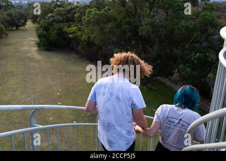 Junges Paar, das davonläuft und die Treppe in einen Stadtpark hinuntergeht. Er hat lockiges orangefarbenes Haar, sie hat blaues Haar. T-Shirt von einem Musikfestival. Perth Stockfoto