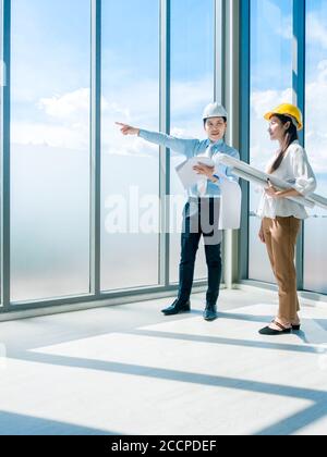 Asiatischer Architekt männlich und weiblich inspizieren Bauarbeiten im Hochhaus in der Nähe von riesigen Glasfenster auf blauem Himmel Hintergrund. Techniker koppeln Stockfoto