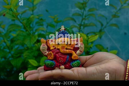 Hölzerne bunte Miniatur lord ganesha Idol auf einer weiblichen Handfläche Mit verschwommener blauer Wand und Baumhintergrund Stockfoto