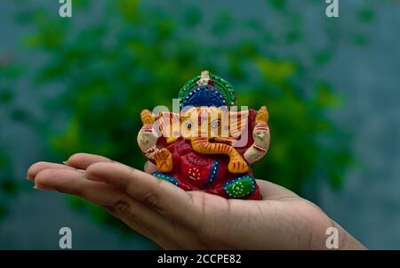 Hölzerne bunte Miniatur lord ganesha Idol auf einer weiblichen Handfläche Mit verschwommener blauer Wand und Baumhintergrund Stockfoto