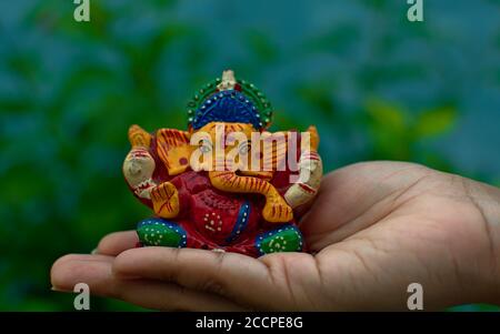 Hölzerne bunte Miniatur lord ganesha Idol auf einer weiblichen Handfläche Mit verschwommener blauer Wand und Baumhintergrund Stockfoto