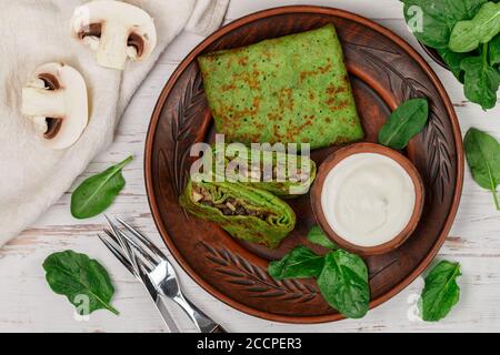 Spinat grüne Pfannkuchen (Crepes) gefüllt mit Pilzfüllung mit saurer Sahne. Köstliches gesundes Frühstück in einem Tonteller auf einem hellen Holzbackgroun Stockfoto