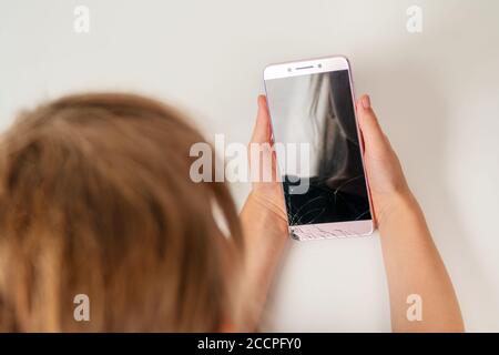 Kind hält ein Smartphone mit einem defekten Bildschirm in seinem Hände und schaut es an Stockfoto