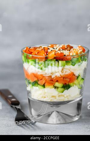 Köstlicher Reissalat mit gesalzenem Lachs (Forelle), Gurke, Avocado, Frischkäse, schwarzen und weißen Sesam in einem Glas auf einem grauen Beton b Stockfoto
