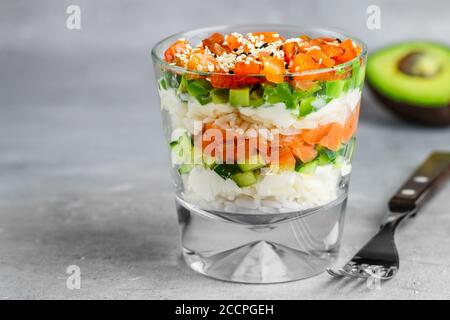 Köstlicher Reissalat mit gesalzenem Lachs (Forelle), Gurke, Avocado, Frischkäse, schwarzen und weißen Sesam in einem Glas auf einem grauen Beton b Stockfoto