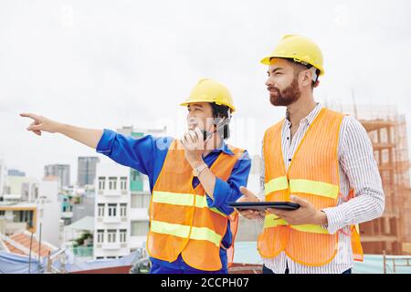 Lächelnder vietnamesischer Vertragspartner, der über Walkie-Talkie spricht und auf den Weg weist, wann Neben dem Chefingenieur mit digitalem Tablet in den Händen Stockfoto