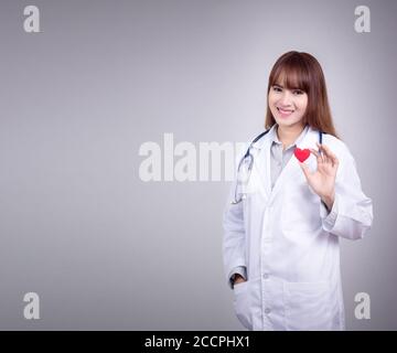 Junge asiatische Arzt stehen mit einem roten Herz in der Hand mit Smiley Gesicht. Dress up mit Arzt Kleid Uniform mit Stethoskop in einem Krankenhaus. Wunderschön Stockfoto