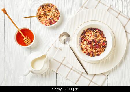 Hafergranola mit getrockneten Himbeeren, Heidelbeeren und Nüssen in einer weißen Schüssel auf einem Holztisch mit Honig und Sahne, Landschaftsansicht von oben, Nahaufnahme, Stockfoto