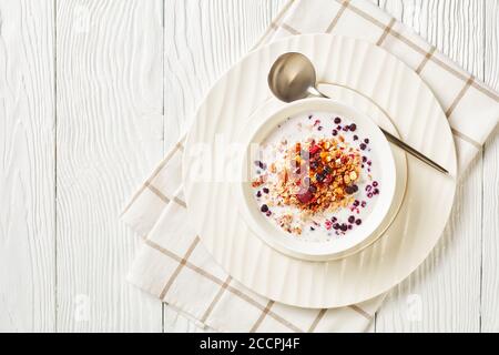 Hafergranola mit getrockneten Himbeeren, Heidelbeeren und Nüssen mit Milch und gekrönt mit Honig in einer weißen Schüssel auf einem Holztisch, Landschaftsansicht von oben, Stockfoto