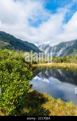 Schneebedeckte Berge spiegeln sich in einem kleinen See an der Westküste Neuseelands. Dunkle Wolken bedecken fast die Gipfel, während gemäßigte Regenwälder die S bedecken Stockfoto