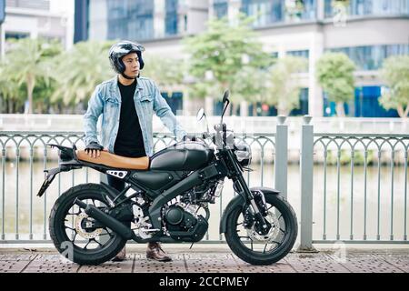 Ernst gut aussehende asiatische Mann im Helm neben seinem stehen Motorrad am Flussufer und wegschauen Stockfoto