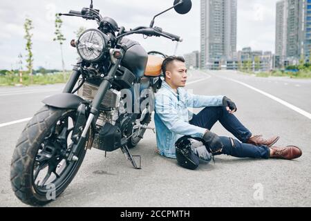 Müde ernsthafte vietnamesische Biker sitzen auf der leeren Straße als nächstes Zu seinem zerbrochenen Motorrad und wegschauen Stockfoto