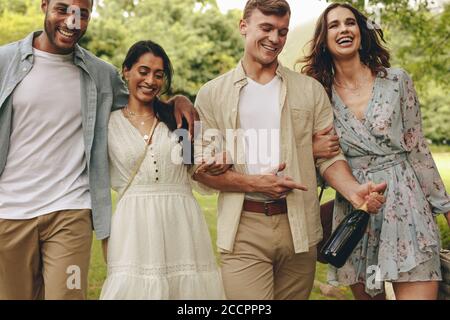 Gruppe junger Leute auf Picknick im Park. Lächelnde Freundinnen und Männer, die mit einer Champagnerflasche durch den Park spazieren. Stockfoto