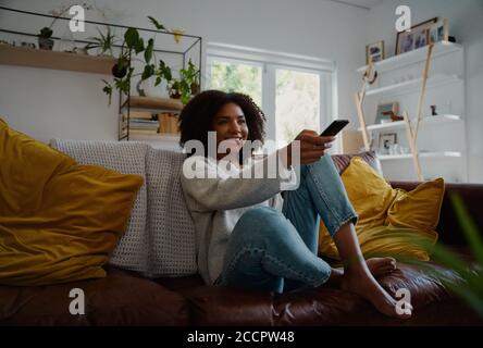 Junge Frau, die drinnen sitzt und fernsieht und die Fernbedienung hält Auf dem Sofa Stockfoto