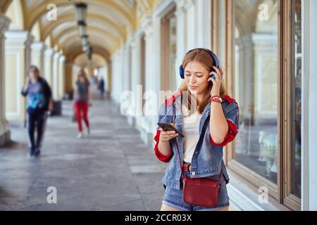 Schöne kaukasische junge fröhliche Frau etwa 25 Jahre alt in Modisches Tuch hört Musik mit Hilfe von Ohrhörern Und Smartphone und Smili Stockfoto