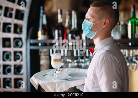 Ein europäisch aussehender Kellner in einer medizinischen Maske serviert Latte-Kaffee. Stockfoto