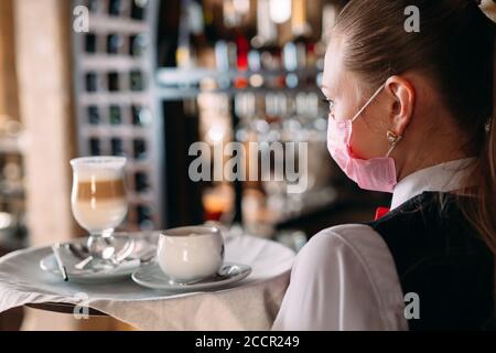 Eine Kellerin mit europäischem Auftritt in einer medizinischen Maske serviert Latte-Kaffee. Stockfoto