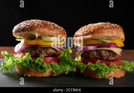 Zwei leckere Burger mit gebratenem Rinderschnitzel, Tomate, Salat und Zwiebeln, knusprigem Weizenmehl-Brötchen mit Sesam. Fastfood auf einem Holzbrett Stockfoto