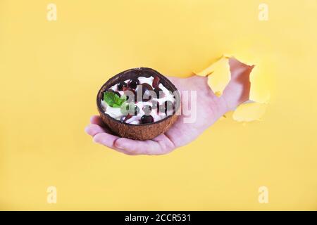 Die Hand eines Mannes in einem Loch hält eine Tasse Kokosnuss mit einem Dessert aus Schlagsahne und Johannisbeeren und Kirschen mit Minzblättern auf gelbem Hintergrund Stockfoto