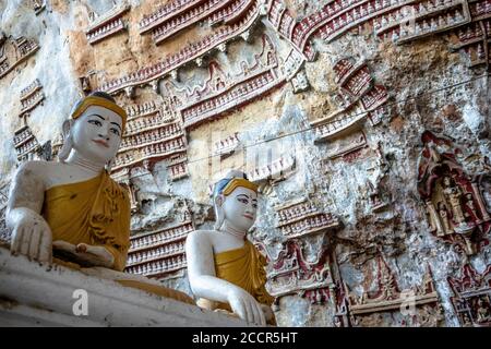 Kleine Figuren, die an den Wänden der Kaw Goon Höhlen geschnitzt wurden. Zwei größere weiße und goldene Statuen, die sitzenden Buddha im Vordergrund darstellen. Hpa An, Myanmar Stockfoto