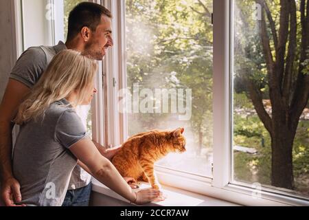 Junges Paar, das am Fenster steht und nach etwas sucht Oder jemand mit ihrer Ingwerkatze in der Nähe Stockfoto