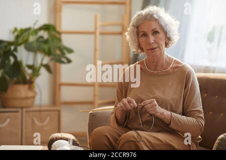 Warm getönte Porträt von eleganten Senior Frau Stricken und Blick auf die Kamera, während auf der Couch sitzen in gemütlichen Hause durch Sonnenlicht beleuchtet, kopieren Raum Stockfoto