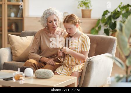 Warm getönte Porträt von eleganten Senior Frau Lehre Enkelin Stricken, während auf der Couch sitzen in gemütlichen Hause durch Sonnenlicht beleuchtet, kopieren Raum Stockfoto