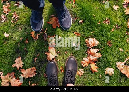 Männliche und weibliche Füße stehen auf dem grünen Gras mit trockenen Herbstblättern, Blick von oben Stockfoto