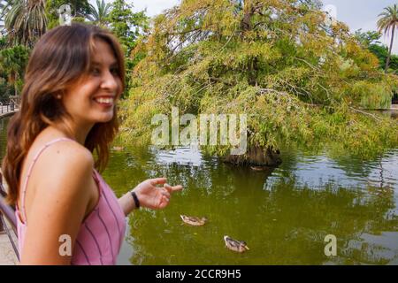 Junge Frau, die um einen lokalen See herum erkundet. Zeigt auf einige Enten im Hintergrund und lächelt. Stockfoto