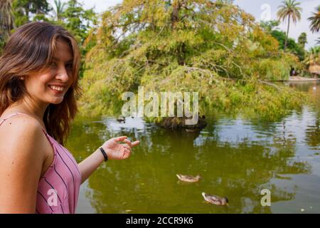 Junge Frau, die um einen lokalen See herum erkundet. Zeigt auf einige Enten im Hintergrund und lächelt. Horizontal. Stockfoto
