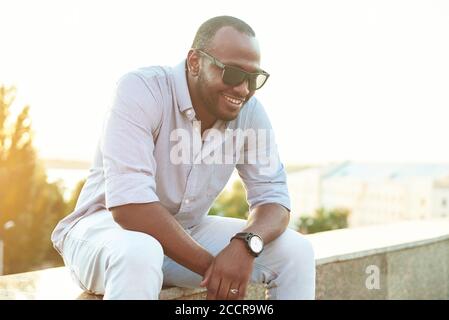 Ein dunkelhäutig Mann mit Sonnenbrille sitzt im Park. Porträt eines schwarzen Mannes aus den späten 20er Jahren, der an einem Sommertag mit gefilterter Wirkung im Park sitzt. Stockfoto