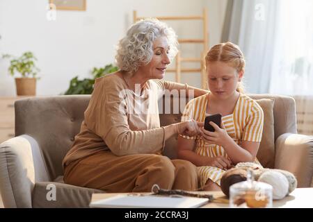 Warm getönte Porträt von eleganten Senior Frau zeigt Smartphone-Fotos Zu niedlichen Enkelin, während sie auf der Couch im gemütlichen Zuhause sitzt Durch Sonnenlicht beleuchtet Stockfoto