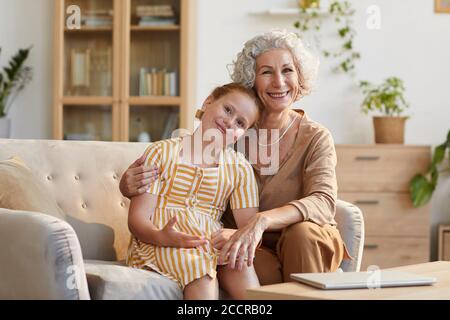 Warm getönte Porträt von lächelnden älteren Frau umarmt niedlich Enkelin, während auf der Couch sitzen und Blick auf die Kamera in gemütlichen Innenraum, kopieren Raum Stockfoto
