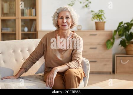 Warm getönte Porträt von eleganten Senior Frau lächelt an der Kamera, während auf der Couch sitzen und Blick auf die Kamera in gemütlichen Innenraum, kopieren Raum Stockfoto