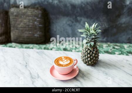 Rosa Tasse Cappuccino und Ananas auf Marmortisch im Loft-Café mit grauer Betonwand und Kissen als Dekoration. Hipster-Style. Stockfoto