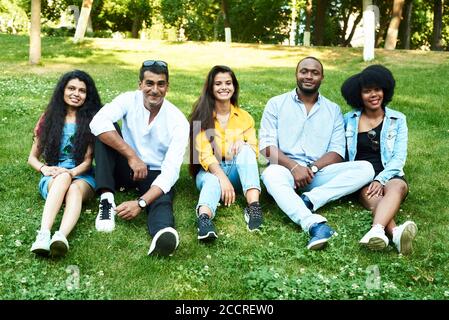 Eine Gruppe von Freunden sitzt auf dem Rasen im Park. Fünf junge Menschen verschiedener Rassen kommunizieren miteinander. Stockfoto