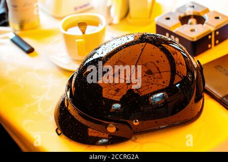 Glänzend schwarzer Motorradhelm im Vintage-Stil mit Regentropfen Auf einem Tisch in einem Café neben einer Tasse Kaffee Stockfoto