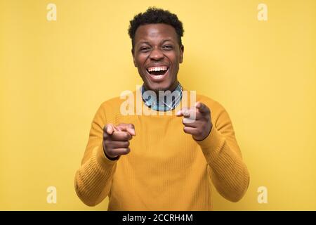 Schöner afrikanischer Mann in gelbem Pullover zeigt auf Kamera auf Witz. Stockfoto