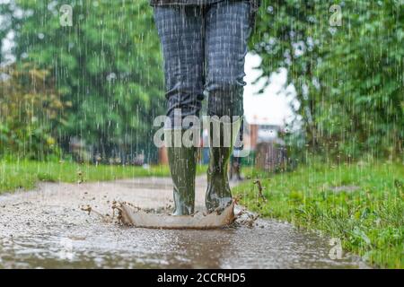 Nahaufnahme, Vorderansicht der grünen Gummistiefel, die von einer Frau getragen werden, die bei starkem Regen in Großbritannien durch Pfützen spritzt. Stockfoto