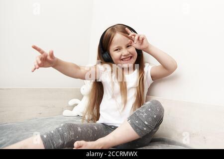 Das Mädchen sitzt in großen Kopfhörern und schaut auf den Smartphone-Bildschirm, sitzt auf ihrem Bett. Vorschulkinder sitzen auf einer Garnstrang, tanzen und singen. Stockfoto